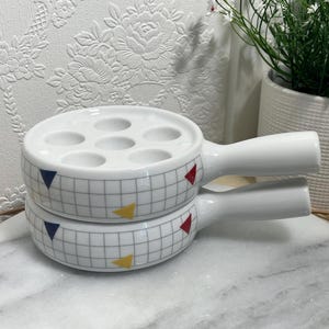 May include: Two stacked white ceramic escargot dishes with handles. Each dish features a grey grid pattern on the exterior, accented with small red, blue, and yellow triangles. The top dish has six circular indentations on its flat surface.