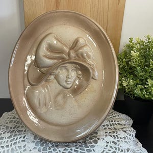May include: Oval ceramic plate featuring a raised relief of a woman in a hat with a bow. The plate is a speckled beige colour with a glossy finish. The woman's face and hat are detailed. Decorative plate.
