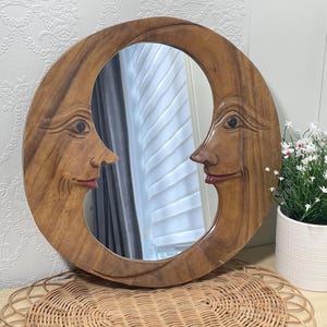 Peut inclure: Un miroir mural circulaire en bois avec un motif sculpté de visage de lune croissante. Le cadre est fabriqué en bois brun avec un grain visible, représentant deux visages de profil se faisant face avec des lèvres peintes en rouge.