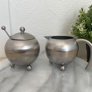 May include: A pair of brushed metal serving pieces: a sugar bowl with a lid and spoon, and a creamer jug with a handle. Both are spherical and sit on three small ball feet. They are on a marble-effect surface, with a small plant in the background.