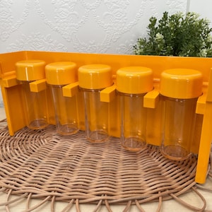May include: A retro spice rack featuring five clear spice jars with orange lids, held in place by an orange plastic holder. The rack is displayed on a woven wicker placemat. The jars are designed to hold spices.