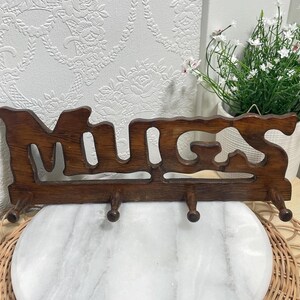 Peut inclure: Un porte-objets mural en bois brun foncé avec le mot MUGS découpé dans une police stylisée. Le support comprend quatre crochets en bois régulièrement espacés s'étendant du bord inférieur, conçus pour suspendre des objets horizontalement.