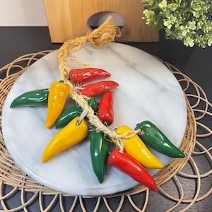 Op de afbeelding: Een decoratieve slinger van keramische chilipepers in rood, geel en groen. De pepers zijn geregen met een natuurlijk vezeltouw en gerangschikt op een wit marmeren serveerplank. De plank rust op een geweven placemat.