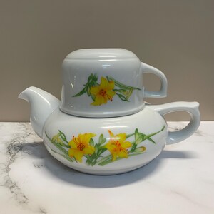Peut inclure: Service à thé en céramique blanche composé d'une théière et d'une tasse. La théière est de forme arrondie avec un bec verseur et une anse. La tasse est posée sur la théière. Les deux pièces sont décorées de fleurs de lys jaunes et de feuilles vertes.