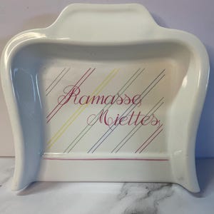Peut inclure: Plateau en céramique blanche avec une poignée et des côtés incurvés. Le plateau porte l'inscription "Ramasse Miettes" en rose, ainsi que des rayures diagonales roses, jaunes, bleues et grises. Un objet décoratif pour la maison.