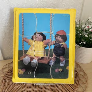 Könnte beinhalten: Ein Vintage-Cabbage-Patch-Kids-Puzzle mit zwei Puppen auf einer Schaukel vor blauem Himmel. Das Puzzle befindet sich in einem gelben Rahmen und scheint sich in der Originalverpackung zu befinden.