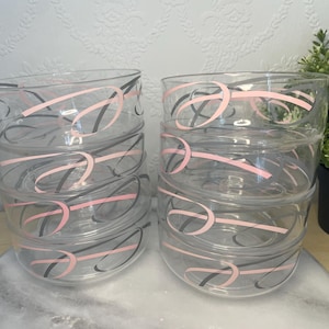 May include: Set of clear glass bowls with pink and black abstract designs. Each bowl is stacked, showcasing the decorative pattern. The bowls are on a marble-like surface, with a potted plant in the background.