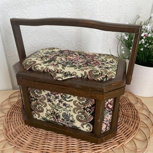 May include: A vintage wooden sewing box with a handle. The hexagonal box features floral fabric on the lid and sides. The fabric has a cream background with pink, green, and brown floral patterns. It rests on a woven placemat.