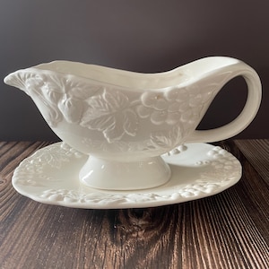 Peut inclure: Saucière en céramique blanche avec sa soucoupe assortie. La saucière est ornée d'un motif en relief de raisins et de feuilles. La soucoupe présente un motif en relief similaire. L'ensemble est posé sur une surface en bois foncé.