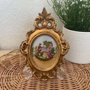 Peut inclure: Cadre doré orné avec un insert ovale en porcelaine représentant une scène pastorale. Le cadre présente un design élaboré et est soutenu par des supports en plastique transparent. L'ensemble dégage une ambiance vintage.