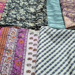 Könnte beinhalten: Eine Sammlung gefalteter Stoffe mit verschiedenen Mustern und Farben, darunter ein dunkelblauer Stoff, ein rosa-orangefarbener geometrischer Druck, ein schwarz-weißes Elefantenmotiv, ein einfarbiges hellblaues Stück und ein violetter Paisleystoff.