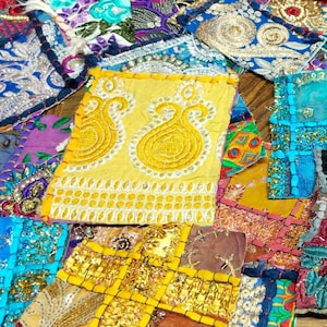 Może przedstawiać: Żywy patchwork z kolorowych kawałków materiału. Projekt obejmuje kwadraty i prostokąty w odcieniach niebieskiego, żółtego, złotego i fioletowego. Niektóre elementy zdobią skomplikowane hafty i koraliki, tworząc teksturowaną i dekoracyjną powierzchnię.