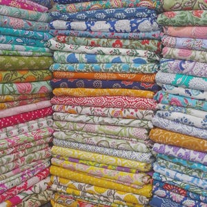 May include: A large collection of folded fabric stacks featuring a variety of colorful block-printed floral and geometric patterns. The textiles display diverse hues including mustard yellow, bright blue, soft pink, sage green, and deep red.