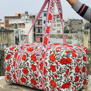 Puede incluir: Un bolso tote rectangular blanco con un estampado floral rojo y hojas verdes. El bolso tiene asas y ribetes a rayas rojas y blancas. El bolso se sostiene frente a un paisaje urbano.