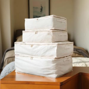 May include: Four off-white storage boxes stacked on a wooden surface. Each box has a zip closure and a fabric handle. The boxes are rectangular and appear to be made of a canvas-like material. The background includes a bed and a framed picture.