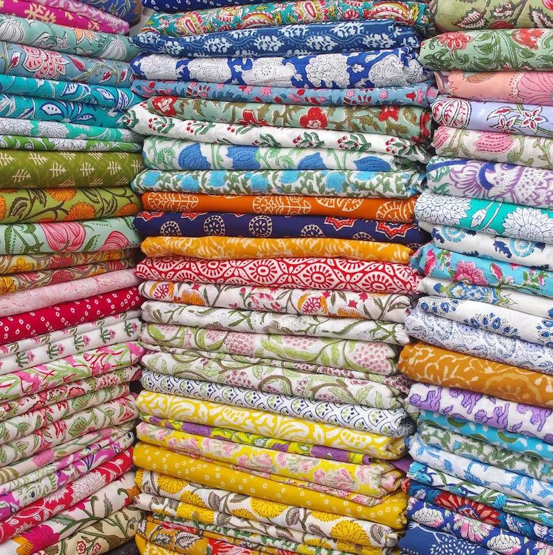 May include: A vibrant display of folded fabric stacks in various colors and patterns. The textiles feature floral, geometric, and paisley designs in shades of blue, green, red, yellow, and pink. The image showcases a diverse collection of textiles.