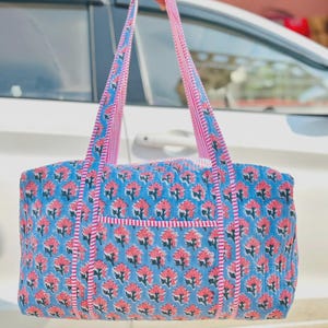 Puede incluir: Un bolso tote azul con un estampado floral rosa. El bolso tiene correas y ribetes rosas. El bolso está hecho de una tela acolchada. El bolso está sostenido por una persona.