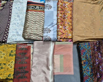 May include: A collection of folded fabric pieces featuring diverse patterns and colors, including brown polka dots, white floral prints, geometric ikat designs, plaid textures, and a vibrant blue and purple textile with intricate border motifs.