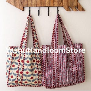 Puede incluir: Dos bolsas de mano con tela estampada, colgadas de ganchos negros en un estante de madera en forma de flecha. Una bolsa tiene un fondo crema con diseños florales azules y rojos. La otra bolsa tiene un patrón de chevron rojo y blanco. El texto "Yashut handloomStore" es visible.