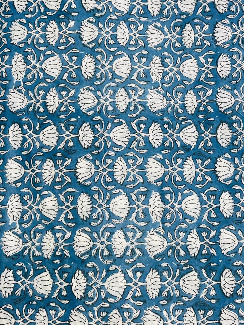 May include: A close-up of a blue fabric with a repeating white floral pattern. The design features stylized flowers and vines, creating a symmetrical and detailed print. The background is a solid, deep blue color.