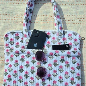 May include: A white quilted tote bag with a pink floral pattern. The bag has two handles and a zipper closure. A black smartphone, sunglasses, and a black card are in the pockets. The bag is on a light-colored surface.
