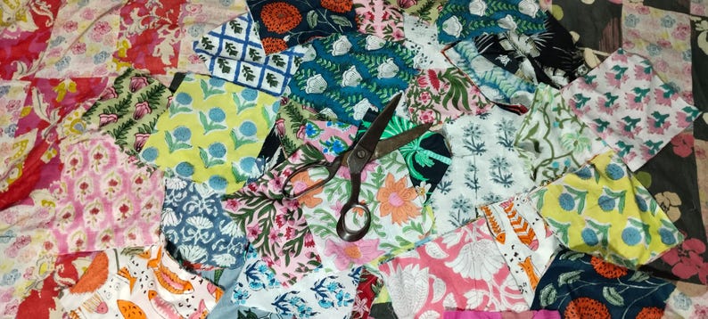 May include: A collection of colorful fabric scraps with various floral and geometric patterns. A pair of vintage scissors rests on top of the fabric pieces. The fabrics feature shades of pink, blue, yellow, and orange.
