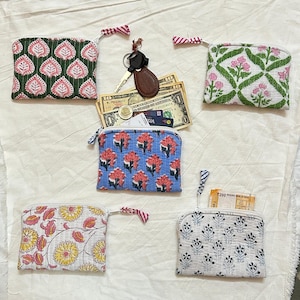 May include: A collection of small, patterned coin purses with zipper closures. The purses feature floral and leaf designs in various colors, including pink, blue, green, and yellow. Some purses are open, revealing cash and a credit card. A set of keys rests nearby.
