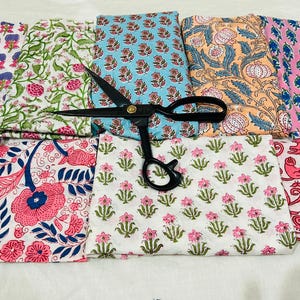 May include: Assortment of colorful fabric swatches with floral and botanical patterns. The fabrics feature pink, blue, green, and orange designs. A pair of black scissors rests on top of the fabric, suggesting a crafting or sewing theme.