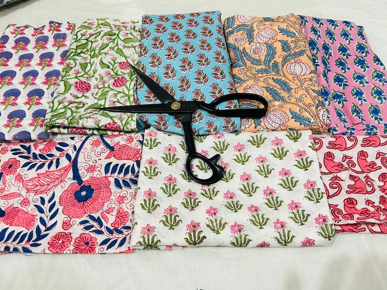 May include: Assortment of colorful fabric swatches with floral and botanical patterns. The fabrics feature pink, blue, green, and orange designs. A pair of black scissors rests on top of the fabric, suggesting a crafting or sewing theme.