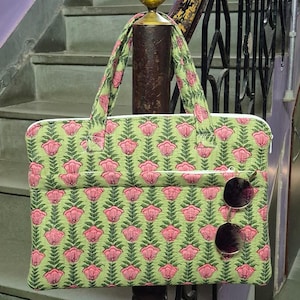 May include: A green and pink floral patterned tote bag with a white zipper and a front pocket. The bag has two handles and a pair of sunglasses hanging from the front. The bag is made of a quilted fabric.