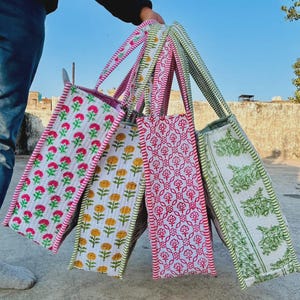Puede incluir: Una colección de bolsas de mano coloridas con estampados florales. Las bolsas presentan varios diseños en rosa, amarillo y verde sobre un fondo blanco. Cada bolsa tiene una correa estampada a juego y un ribete rosa o verde contrastante.