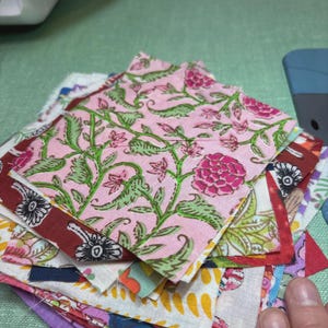 May include: A stack of colorful fabric squares, featuring floral patterns in pink, green, and burgundy. The squares are of various sizes and designs, with a focus on a pink floral print. The fabric appears to be cotton or a similar material.