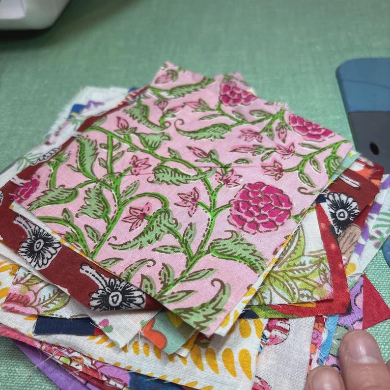 May include: A stack of colorful fabric squares, featuring floral patterns in pink, green, and burgundy. The squares are of various sizes and designs, with a focus on a pink floral print. The fabric appears to be cotton or a similar material.