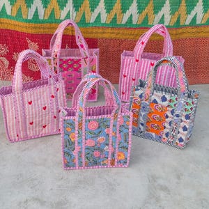 Op de afbeelding: Vijf handgemaakte tote tassen in verschillende maten en patronen. De tassen hebben roze en witte gestreepte stof, bloemenprints en rode hartaccenten. Elke tas heeft een stevige handgreep en een gewatteerd ontwerp.