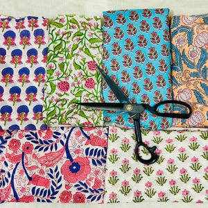 May include: Assortment of colorful floral patterned fabrics in various designs and colors, including pink, blue, green, and orange. A pair of black scissors is placed on top of the fabrics, suggesting a craft or sewing project.