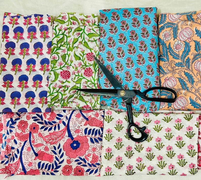 May include: Assortment of colorful floral patterned fabrics in various designs and colors, including pink, blue, green, and orange. A pair of black scissors is placed on top of the fabrics, suggesting a craft or sewing project.