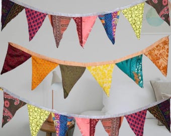 May include: A colorful fabric banner with triangular flags in various patterns and colors, including pink, orange, blue, and green. The banner is strung on a white ribbon, creating a festive and decorative element.