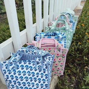 May include: A collection of quilted tote bags in various floral and striped patterns. The bags feature blue, pink, and green designs with white accents. The bags are arranged on a white fence, with greenery in the background.