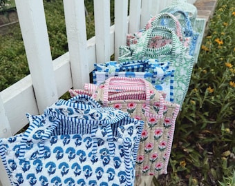 May include: A collection of quilted tote bags in various floral and striped patterns. The bags feature blue, pink, and green designs with white accents. The bags are arranged on a white fence, with greenery in the background.