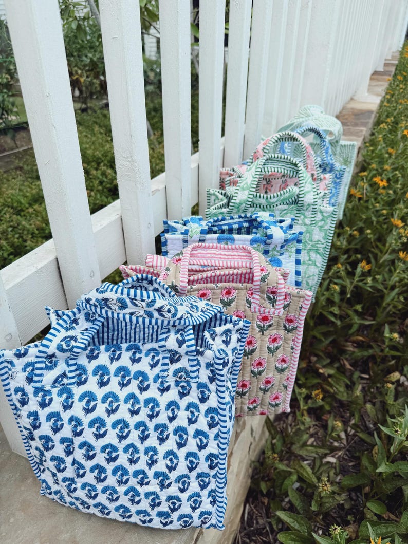 May include: A collection of quilted tote bags in various floral and striped patterns. The bags feature blue, pink, and green designs with white accents. The bags are arranged on a white fence, with greenery in the background.
