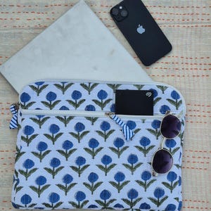 Könnte beinhalten: Eine weiße Laptoptasche mit blauem Blumenmuster, ein schwarzes Smartphone, eine Sonnenbrille und eine schwarze externe Festplatte. Die Tasche hat einen Reißverschluss und blau-weiß gestreifte Reißverschlussanhänger. Der Hintergrund ist ein cremefarbener Stoff.