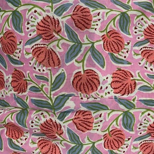 May include: Pink fabric with a repeating floral pattern. The design features large, coral-colored flowers with detailed petals, blue-gray leaves, and green stems. The background is a soft pink.