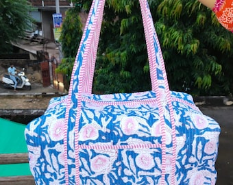 Pode incluir: Uma sacola tote com estampa floral azul e branca com detalhes em rosa. A sacola tem formato retangular com duas alças e um bolso frontal. As alças e bordas são aparadas em rosa. A sacola é segurada por uma pessoa.