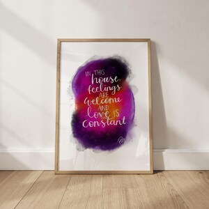 May include: A framed art print with a watercolor design. The artwork features a vibrant purple and pink abstract shape with the words "IN THIS house, feelings are welcome AND love IS constant" in white script.