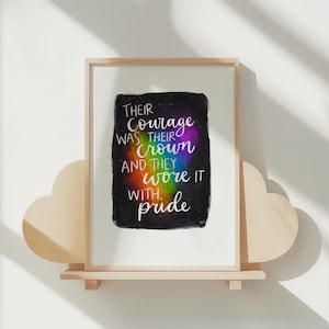 Può includere: Stampa artistica incorniciata con il testo "Their courage was their crown and they wore it with pride." L'opera d'arte presenta uno sfondo nero con un centro dai colori dell'arcobaleno. La cornice è in legno chiaro e la stampa è esposta su una mensola a forma di nuvola.