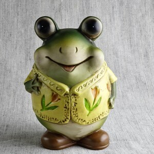 以下が含まれることがあります： 大きな目と愛想の良い笑顔が特徴的な、セラミック製のカエルの置物。カエルは、花の模様が施された黄色のベストを着ています。肌は緑とクリーム色で、茶色の靴を履いています。