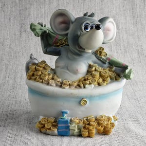 May include: A whimsical figurine of a gray mouse relaxing in a bathtub filled with gold coins. The mouse holds bags of money and is surrounded by more coins. The bathtub is white and blue, with additional coins and soap bottles at the base.