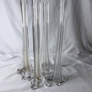 Peut inclure: Un ensemble de quatre vases soliflores en verre transparent, hauts et élancés, au design cannelé. Chaque vase présente une base évasée en forme d'étoile et des stries verticales le long du corps, se rétrécissant vers une ouverture étroite.