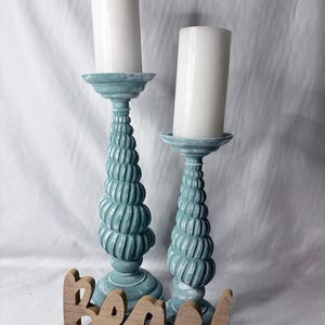 Peut inclure: Deux bougeoirs sarcelle avec une texture en spirale rappelant un coquillage et une finition vieillie, surmontés chacun d'une bougie pilier blanche. Devant les bougeoirs se trouve une inscription en bois découpé avec le mot beach en cursif.