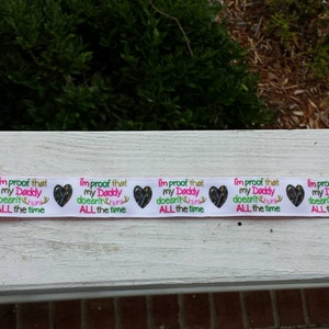 May include: White fabric baby clip with a pink and green patterned design. The clip has a metal clasp and a white ribbon with the text "I'm proof that my Daddy doesn't hunt ALL the time" repeated five times.